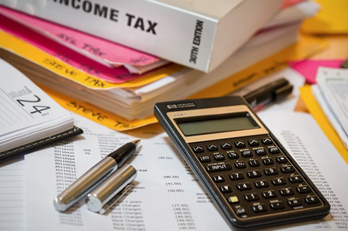 A calculator, pens, and documents are spread across a table in the Seattle Public Library. A large book labeled "Income Tax" is partially visible in the background, hinting at a busy session of Seattle tax services or tax prep underway.