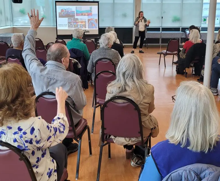 In a membership meeting, a group of people seated in a room listens intently to the speaker near the presentation screen. One person raises a hand, likely for a question or to ask for a quick recap.