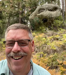 Rock hunting enthusiast Dave, sporting glasses and a mustache, smiles for a selfie in front of a large mossy rock with holes, surrounded by forest trees and plants.