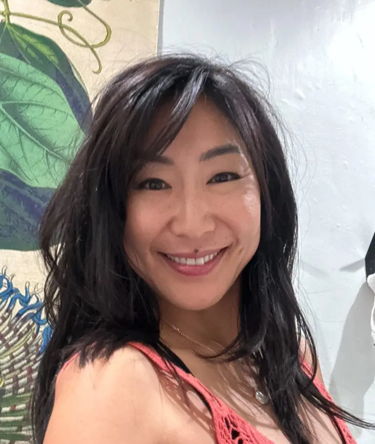 Photo of Nally Berg, a woman with long dark hair smiles at the camera, standing indoors near a wall with a large green leaf design