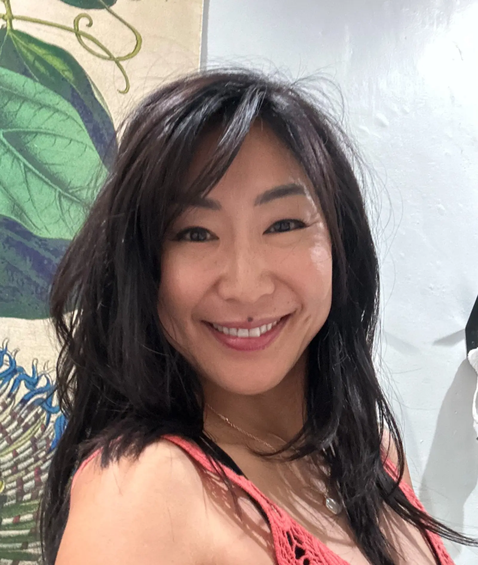 Photo of Nally Berg, a woman with long dark hair smiles at the camera, standing indoors near a wall with a large green leaf design
