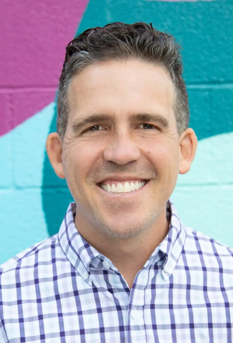 Christopher with short dark hair, a board staff member, smiles at the camera, wearing a plaid shirt, standing in front of a colorful mural with purple, teal, and light blue shapes.