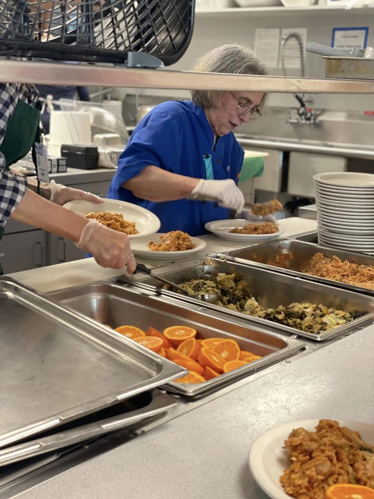 Two people in a cafeteria kitchen serve food onto plates. Various food items, including oranges, are displayed in trays. The woman in a blue shirt and white gloves is scooping food from a tray, embodying the essence of food and dining at The Center for Active Living.
