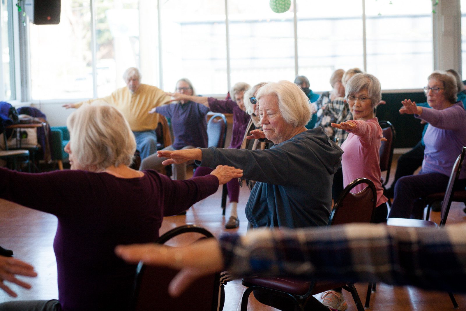 A group of elderly individuals participate in a seated exercise class, extending their arms forward while seated in a well-lit room.
