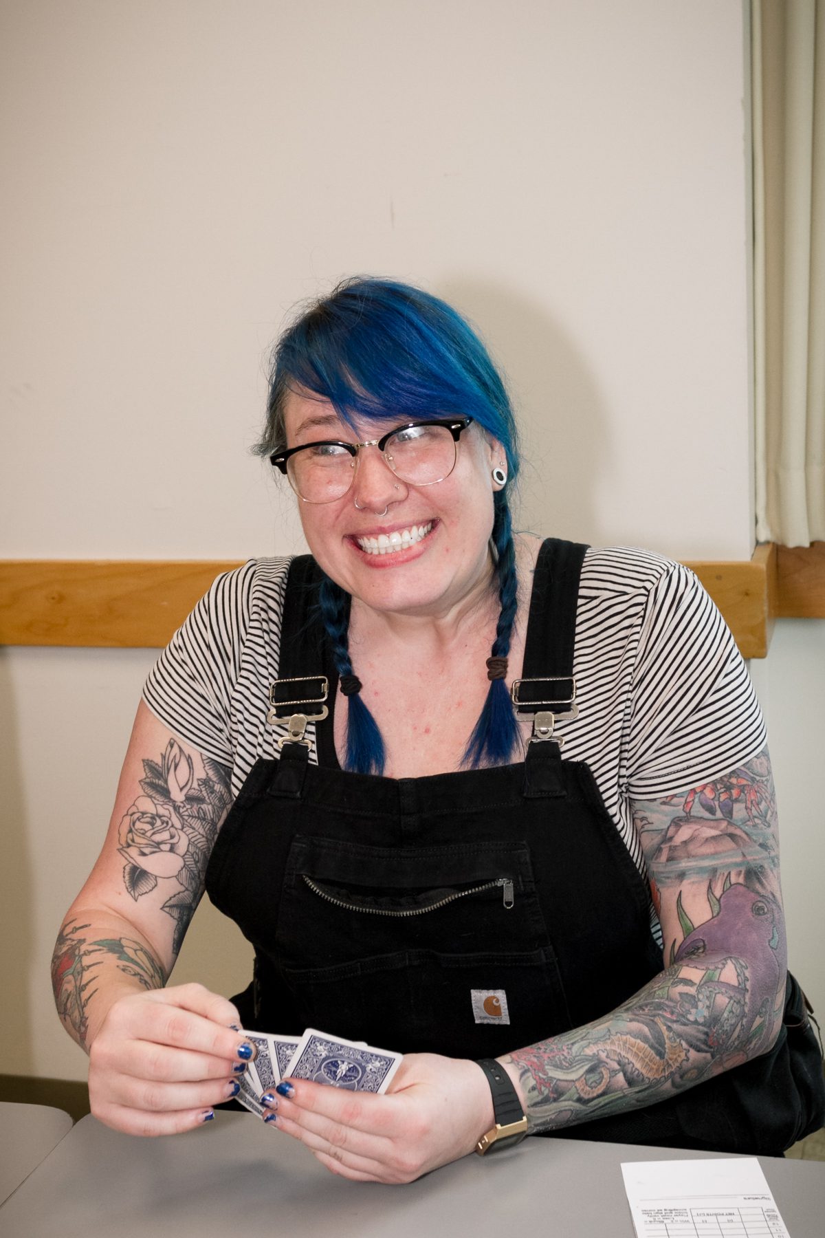 A person with blue hair, glasses, and tattoos, wearing a striped shirt and overalls, is sitting at a table holding playing cards.
