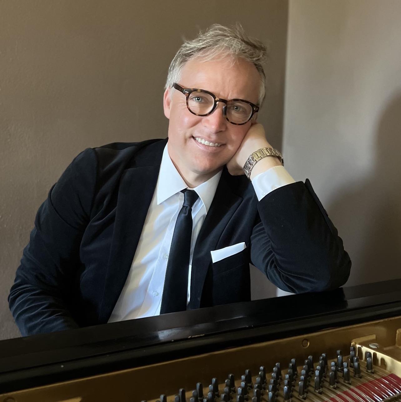 Larry Knapp, dressed in a suit and tie, sits at a piano and smiles while resting their head on their hand.