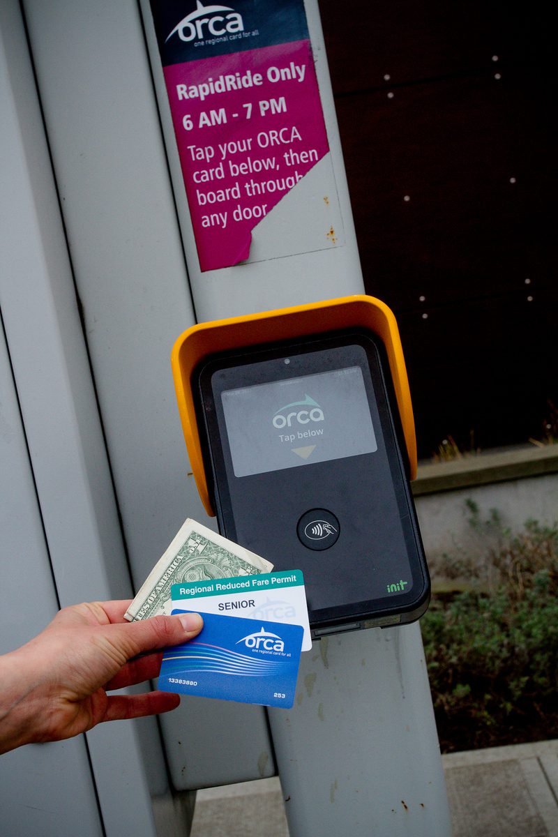 Person holding a $1 bill, a Regional Reduced Fare Permit card, and an ORCA card next to an ORCA card reader at a bus station, demonstrating the transportation help available at The Center for Active Living.