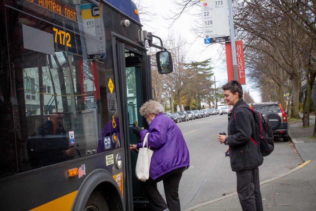 A woman wearing a purple jacket boards a city bus, while a man with a backpack waits behind her. A bus stop sign and parked cars are seen along the street, perhaps hinting at the transportation help available at The Center for Active Living nearby.