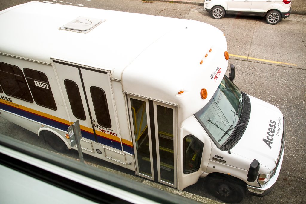 A white Access Metro shuttle bus, providing transportation help at The Center for Active Living, is parked on the street next to a sidewalk. It features a curved roof and a front door that is slightly open. A white SUV is parked in front of it.