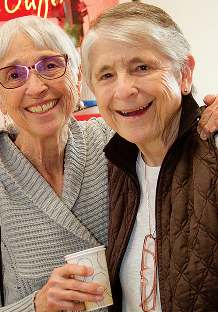 Two elderly women smiling in the final photo: one with glasses and a gray sweater holding a cup, the other wearing a dark vest with reading glasses hanging from her neck.