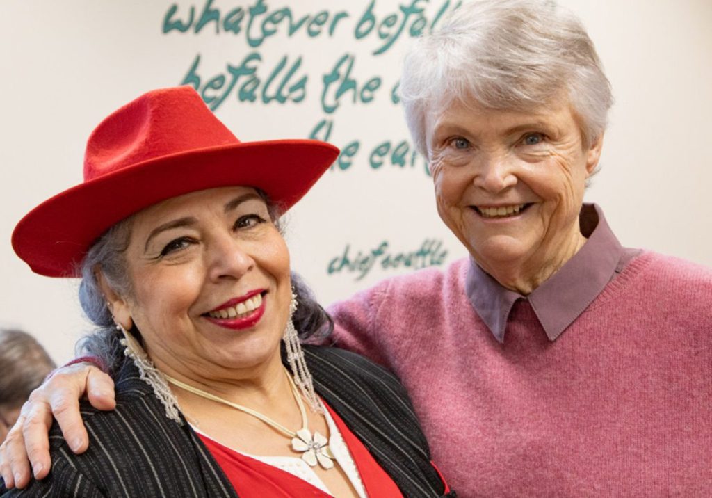 Two smiling women posing together; one wears a red hat and black jacket with a white shirt, and the other wears a pink sweater over a collared shirt. The background features a partial text quote on the wall, captured perfectly in these FINAL GLOBAL PHOTOS.