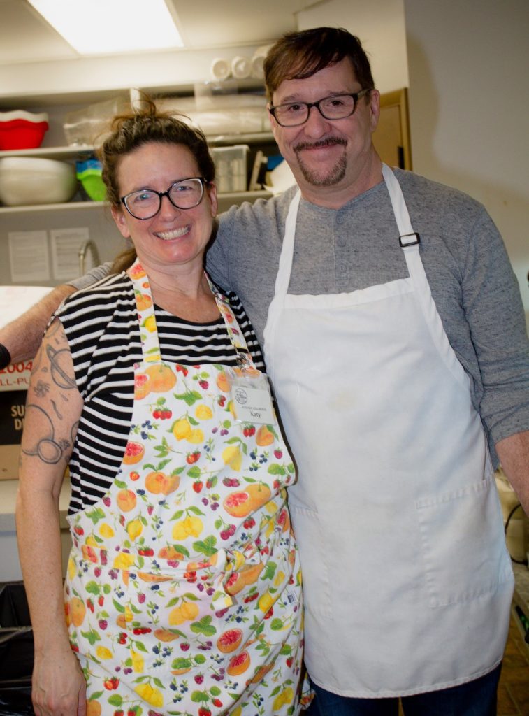 Two individuals wearing aprons stand close together in a kitchen, smiling at the camera. The person on the left wears a colorful, fruit-patterned apron, while the person on the right is in a plain white apron. Get involved at The Center for Active Living to join in on fun activities like this!