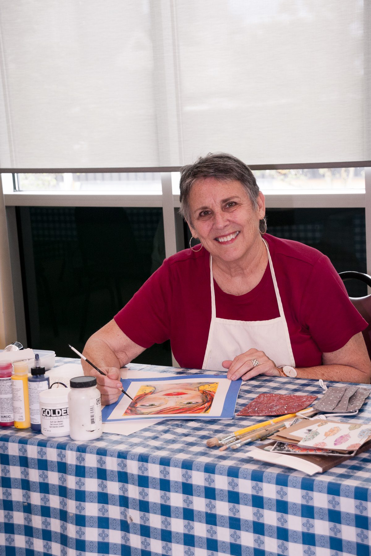 An older woman, embodying the spirit of lifelong learning, is seated at a table with painting supplies. Wearing a red shirt and apron, she smiles while working on a painting at The Center for Active Living West Seattle.