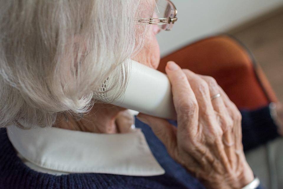 An elderly woman with white hair and glasses holds a landline telephone to her ear while sitting on a chair, reaching out to The Center for Active Living for counseling and support groups.