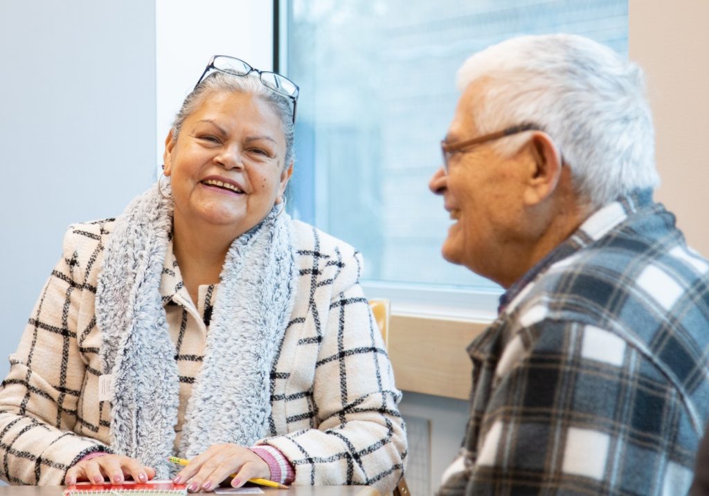 Two older adults sit at a table by a window, smiling and talking. One woman wears glasses, a grey scarf, and a checkered jacket; the man wears glasses and a plaid shirt.