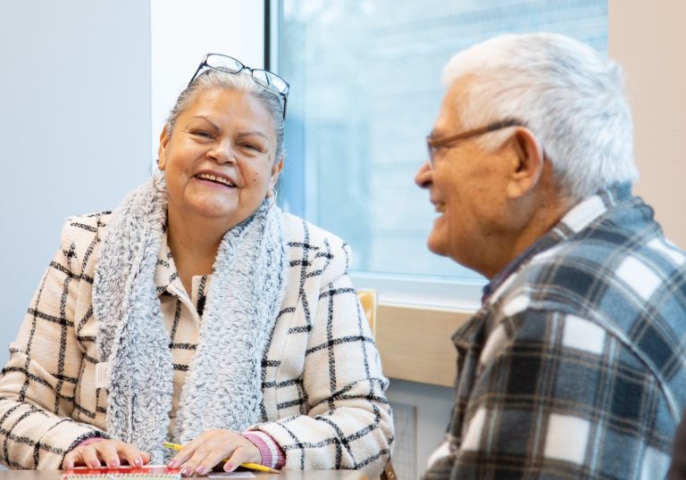 Two older adults sit at a table by a window, smiling and talking. One woman wears glasses, a grey scarf, and a checkered jacket; the man wears glasses and a plaid shirt.