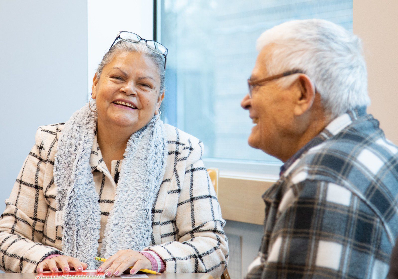 Two older adults sit at a table by a window, smiling and talking. One woman wears glasses, a grey scarf, and a checkered jacket; the man wears glasses and a plaid shirt.