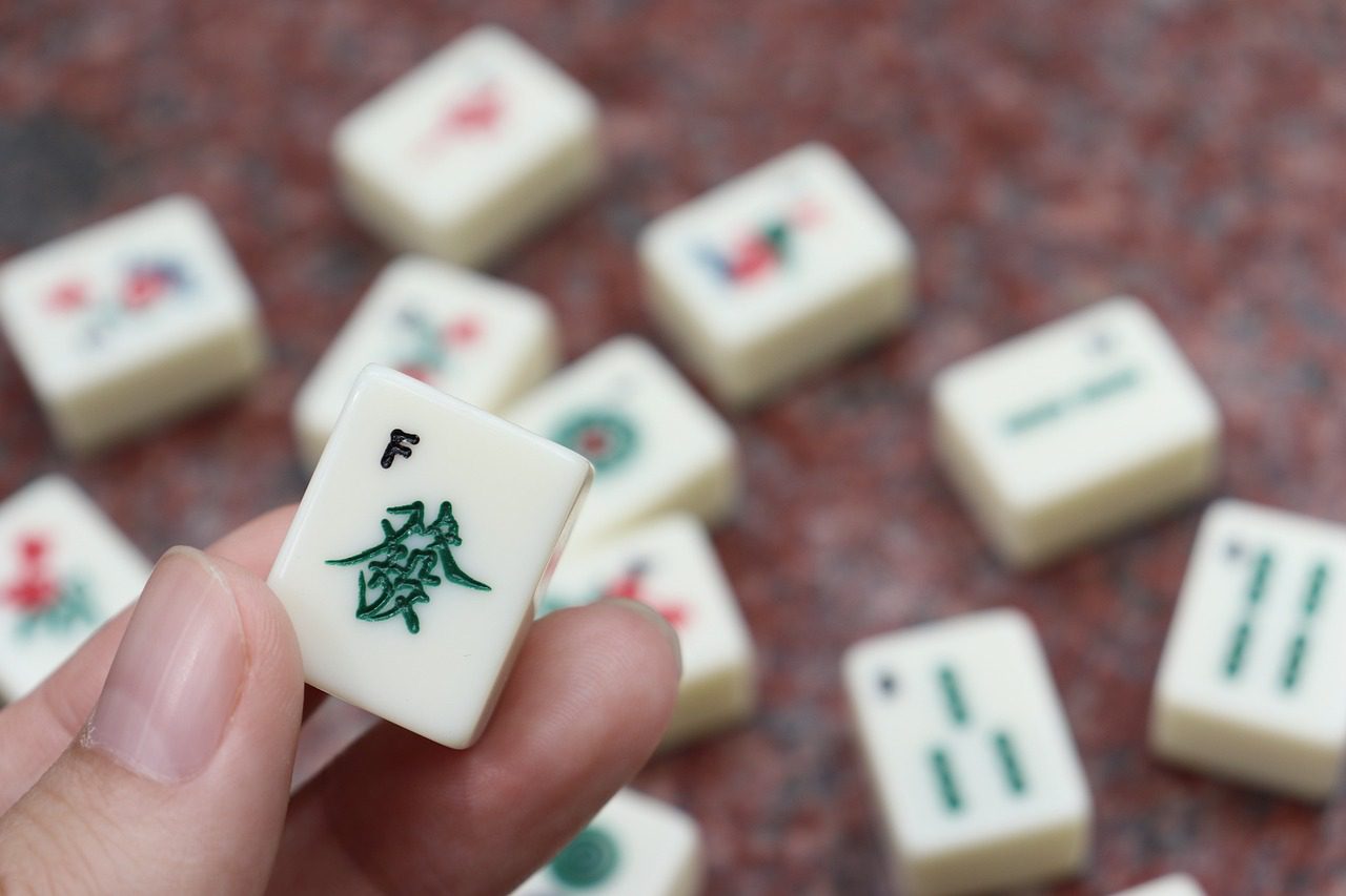 A hand holding a single Mahjong tile, with multiple other tiles scattered on a flat surface in the background, reminiscent of an American Mahjong game at The Center for Active Living West Seattle.