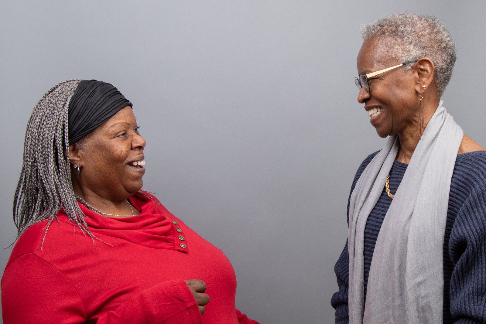 Two women stand facing each other, smiling and laughing, against a plain gray background.