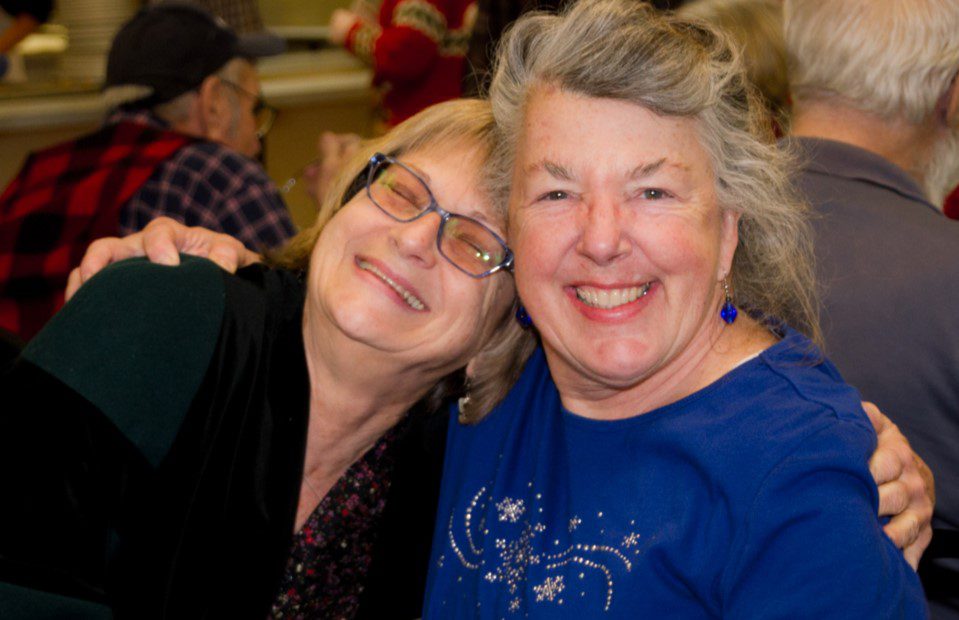 Two women smiling, one wearing glasses and a floral top, the other in a blue shirt. They are closely leaning into each other in a busy indoor setting, showing the warm connections you can make when you get involved at The Center for Active Living.