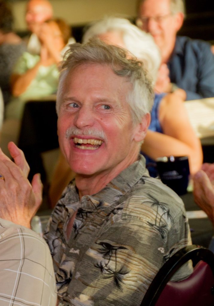 A man with gray hair and a mustache smiles while clapping. He wears a patterned shirt and is surrounded by other people in a well-lit indoor setting. For more moments like these, contact The Center for Active Living.