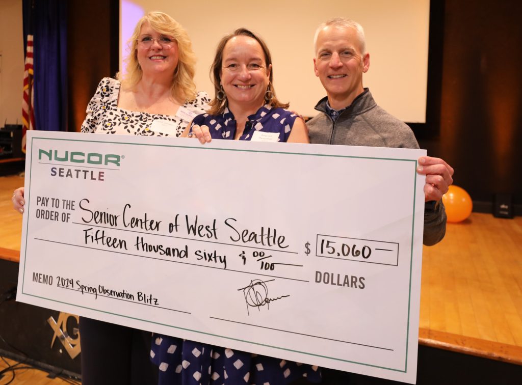Three people smiling and holding a large ceremonial check for $15,060 made out to the Senior Center of West Seattle from Nucor Seattle, on stage in an indoor setting during the Annual Fundraising Breakfast for The Center for Active Living.