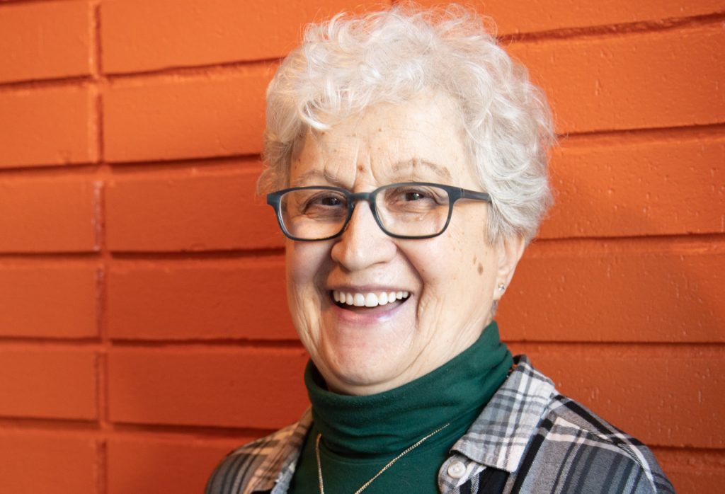 An elderly woman with short gray hair and glasses smiles in front of an orange brick wall. She is wearing a green turtleneck and a plaid shirt. For more senior activities, contact The Center for Active Living.