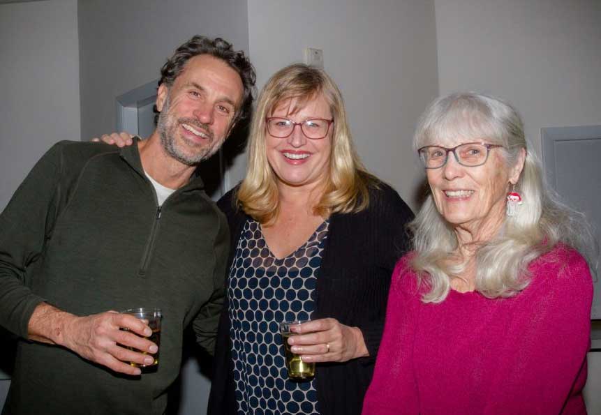Three people are standing indoors, smiling at the camera. The two women are holding drinks, while the man, who's just been discussing great activities for seniors in West Seattle, is holding a drink in one hand and has his other arm around one of the women.