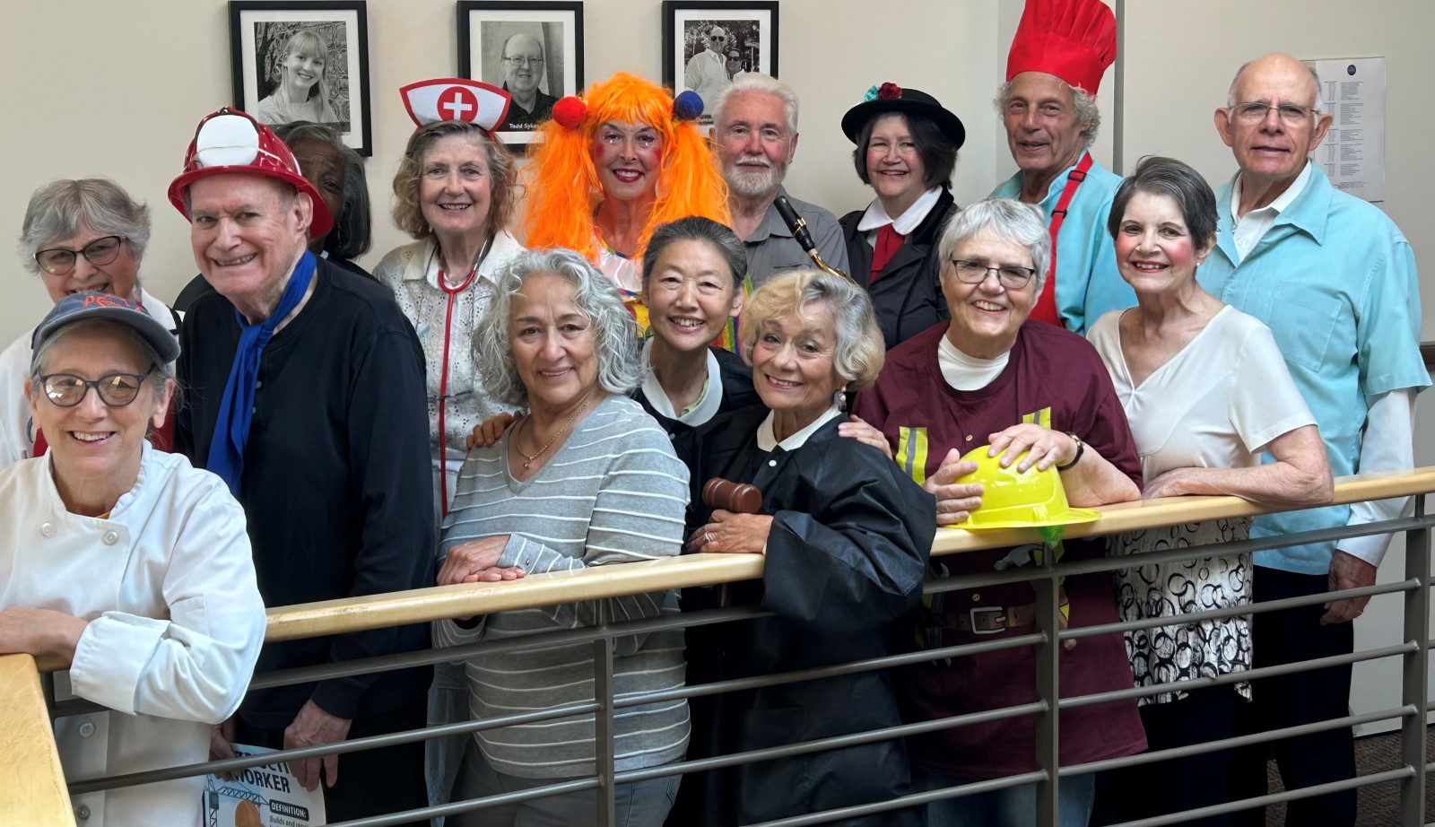 Group of Silver Sounds performers from West Seattle pose indoors, many wearing festive costumes such as a chef, nurse, clown, and judge, smiling near the railing