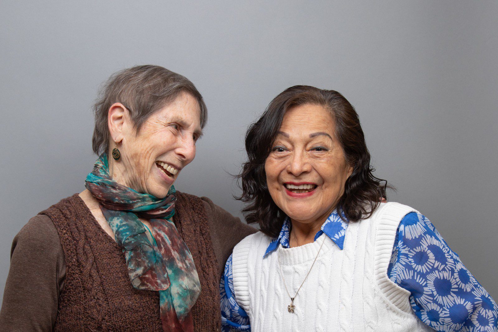 Two women stand close together, smiling and laughing in front of a plain gray background.