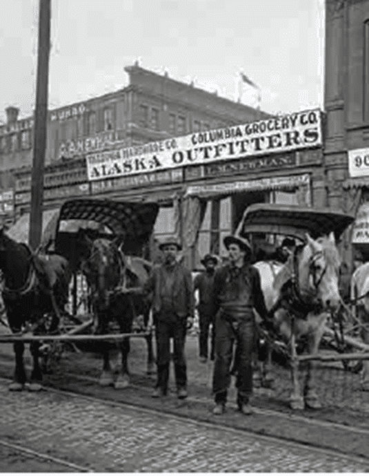 Two men stand beside horse-drawn wagons on a cobblestone street, in front of buildings with signs for Columbia Grocery Co. and Alaska Outfitters—echoes of the Klondike stampede and gold fever linger in the air.