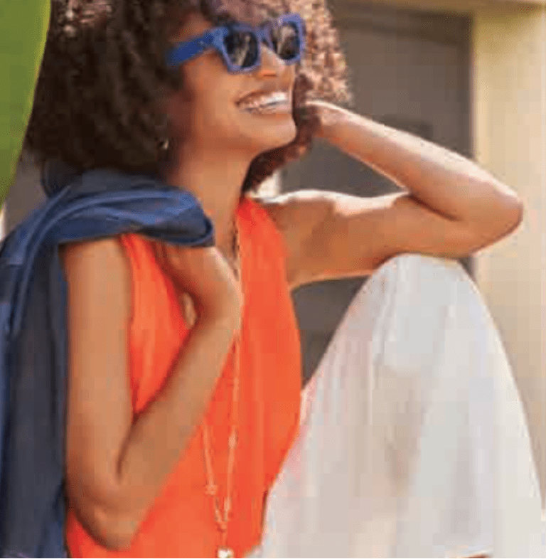 A woman with curly hair wearing sunglasses, an orange sleeveless top, and white pants sits outdoors smiling, a blue scarf draped over her shoulder