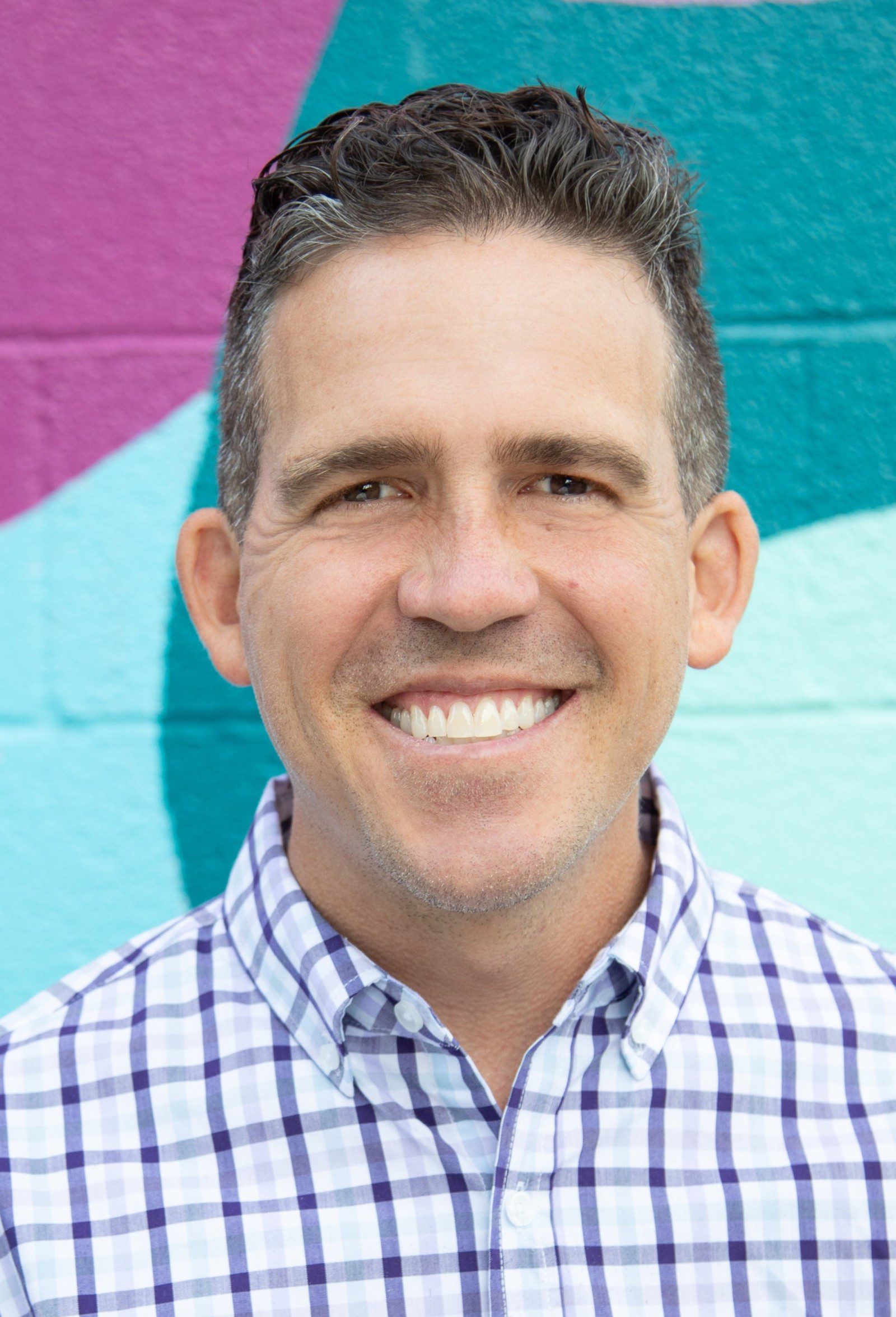 Christopher with short dark hair, a board staff member, smiles at the camera, wearing a plaid shirt, standing in front of a colorful mural with purple, teal, and light blue shapes.