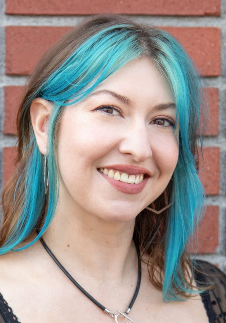 Yoni with blue-dyed hair and hoop earrings stands in front of a brick wall, looking like a vibrant staff member.