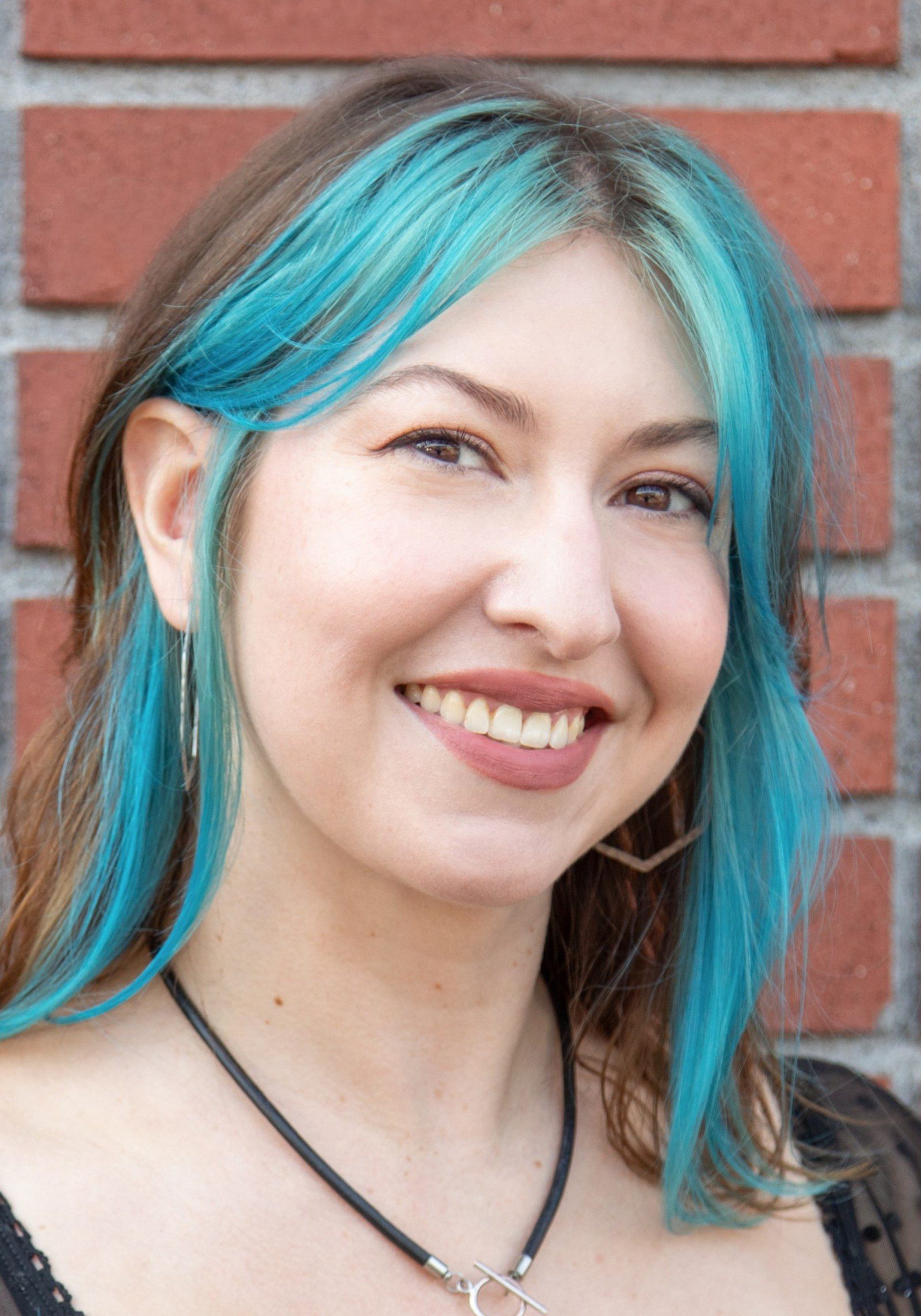 Yoni with blue-dyed hair and hoop earrings stands in front of a brick wall, looking like a vibrant staff member.