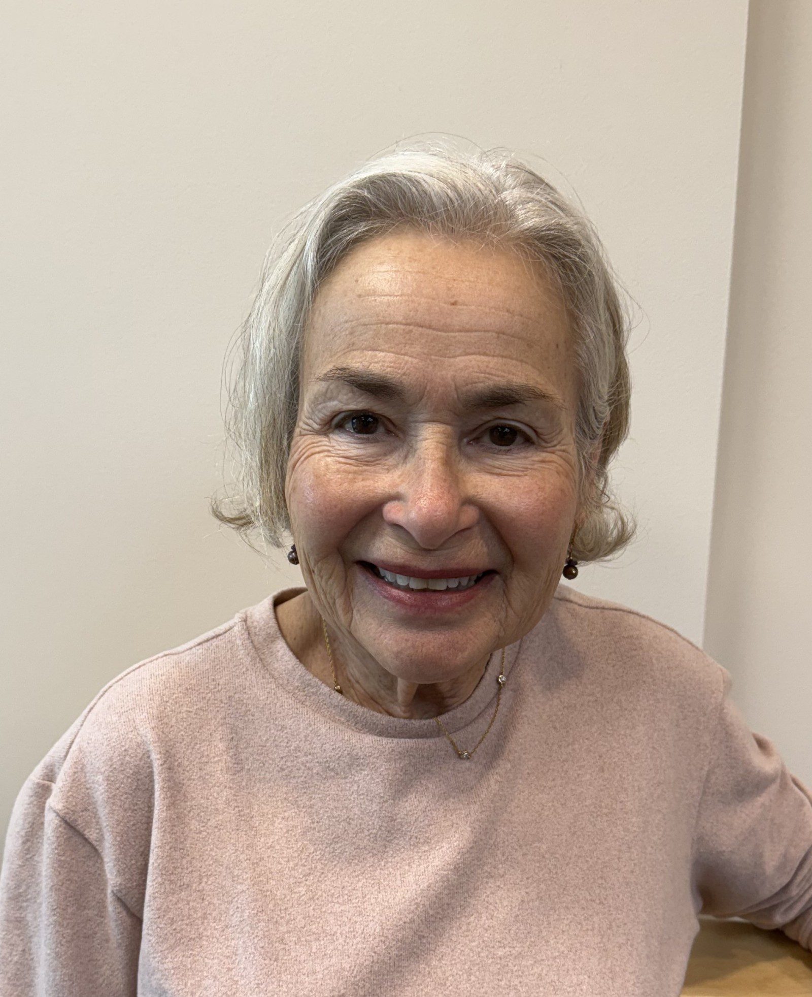 Photo of Geri, who has gray hair and is smiling at the camera, wearing a light pink sweater and simple jewelry