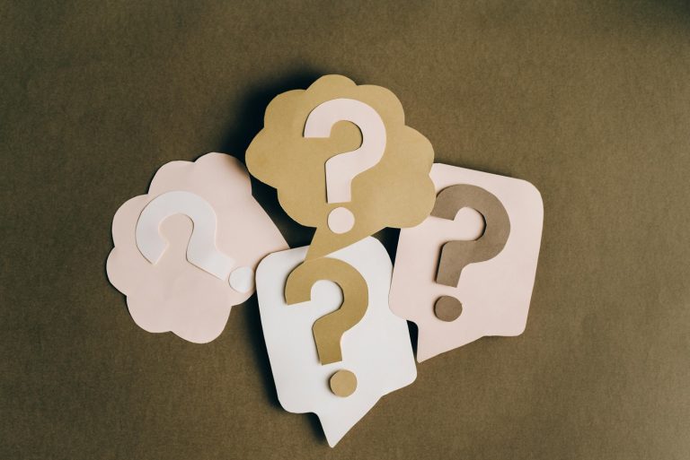 Four paper speech bubbles in beige and white, each with a question mark cut out, are arranged on a brown surface.