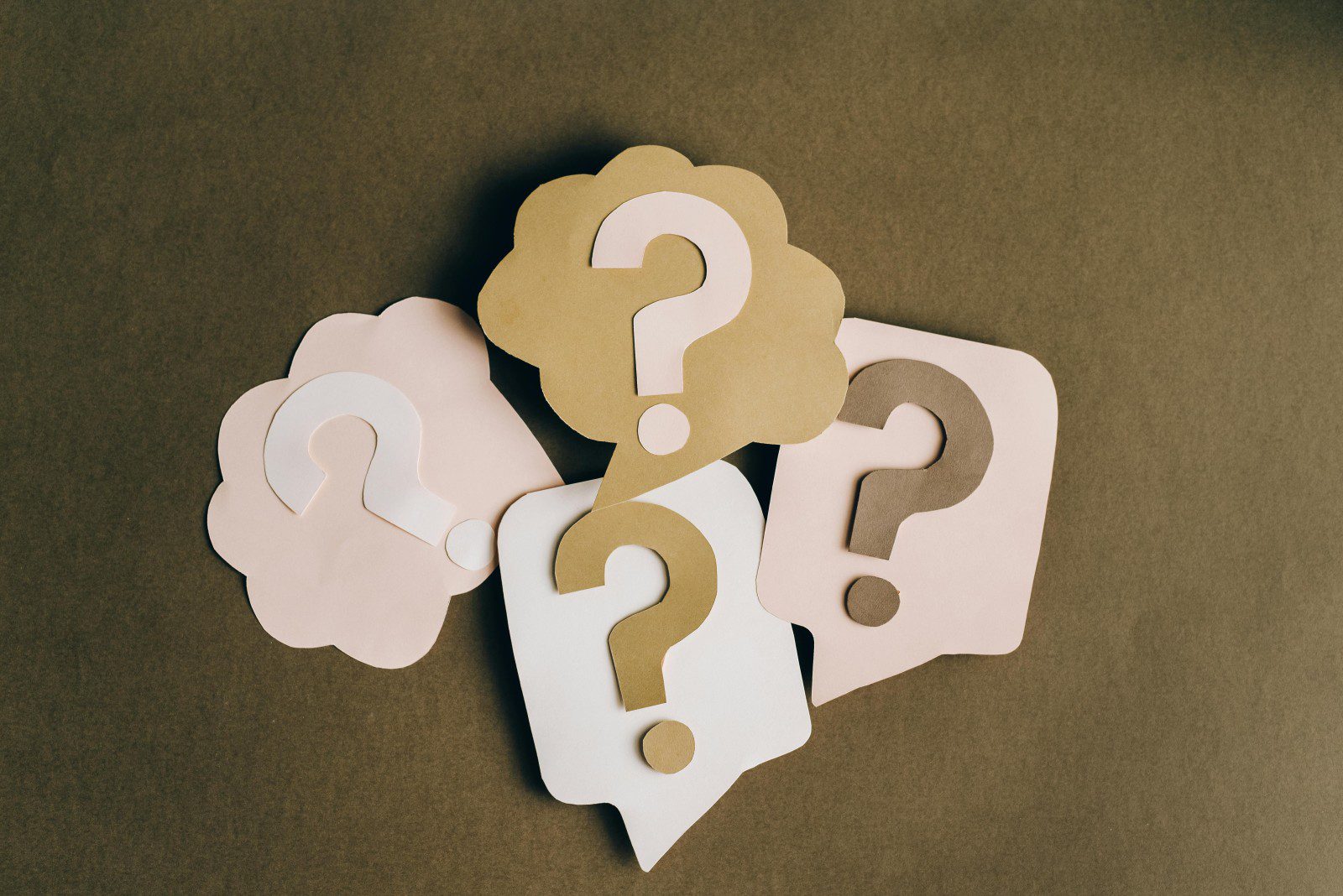 Four paper speech bubbles in beige and white, each with a question mark cut out, are arranged on a brown surface.