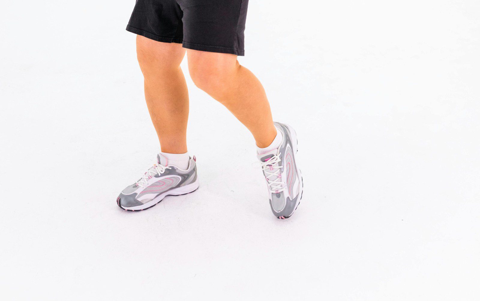 Person wearing black shorts and gray sneakers, standing on a white background with legs slightly crossed.