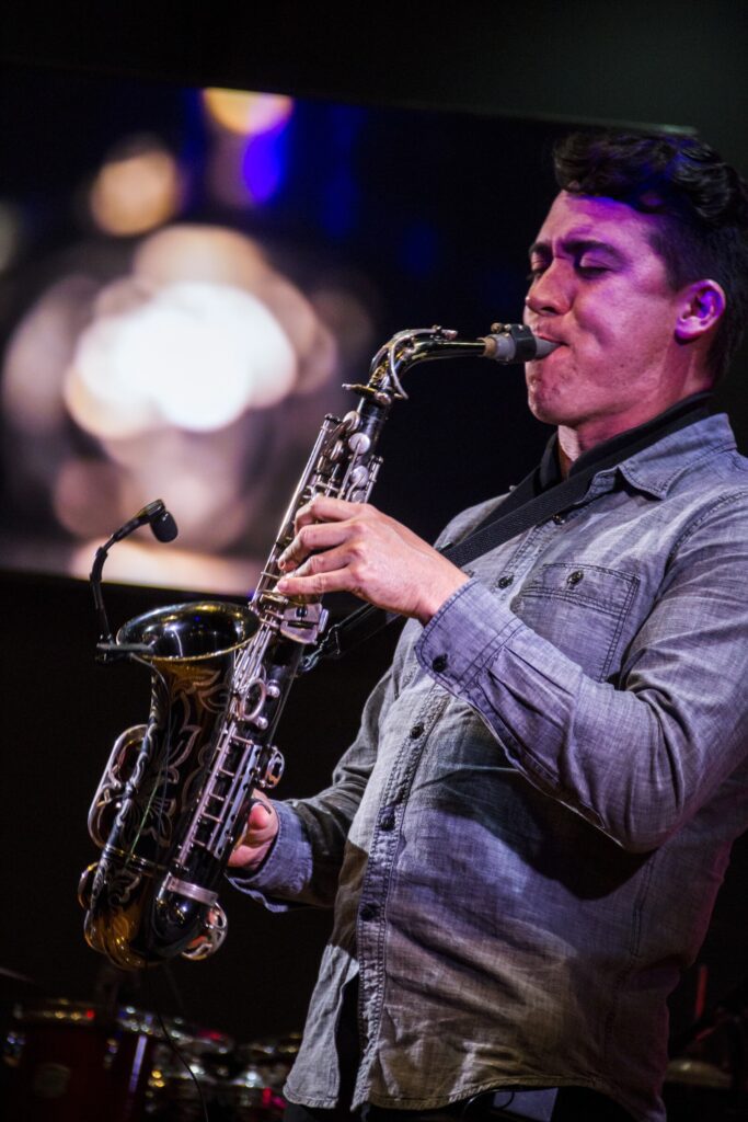 A man in a gray shirt plays a saxophone on stage, enveloped by blurred lights and a dark background, capturing the soulful energy of jazz.