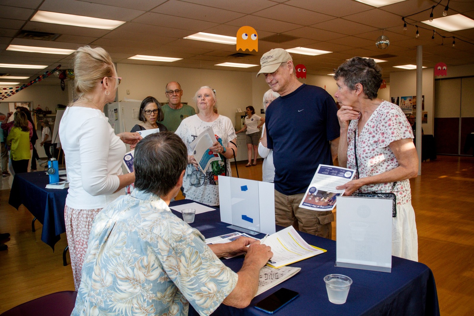 People stand at a table speaking with two staff members at an indoor event, with informational materials and decorations visible in the room