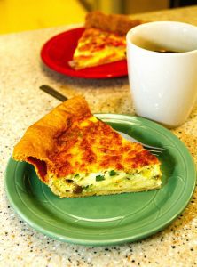 A slice of quiche on a green plate with a fork, next to a white mug of black coffee; another slice is on a red plate in the background, creating an inviting dining scene for anyone who appreciates good food.
