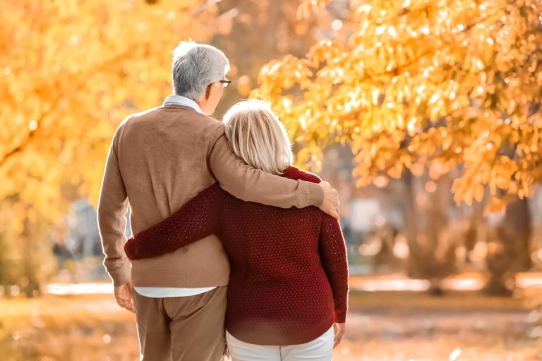 An older couple walks outdoors in a park during autumn, with yellow leaves on trees.