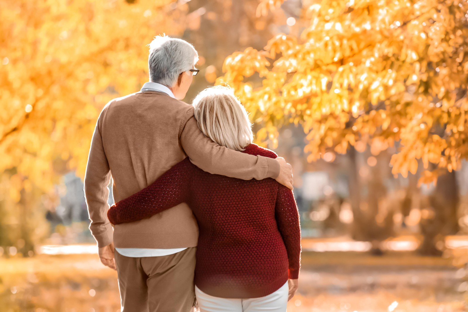 An older couple walks outdoors in a park during autumn, with yellow leaves on trees.