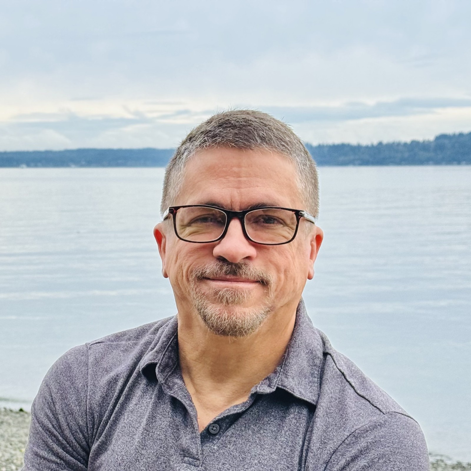 Headshot of David with short gray hair, glasses, and a trimmed beard sits outdoors near a body of water