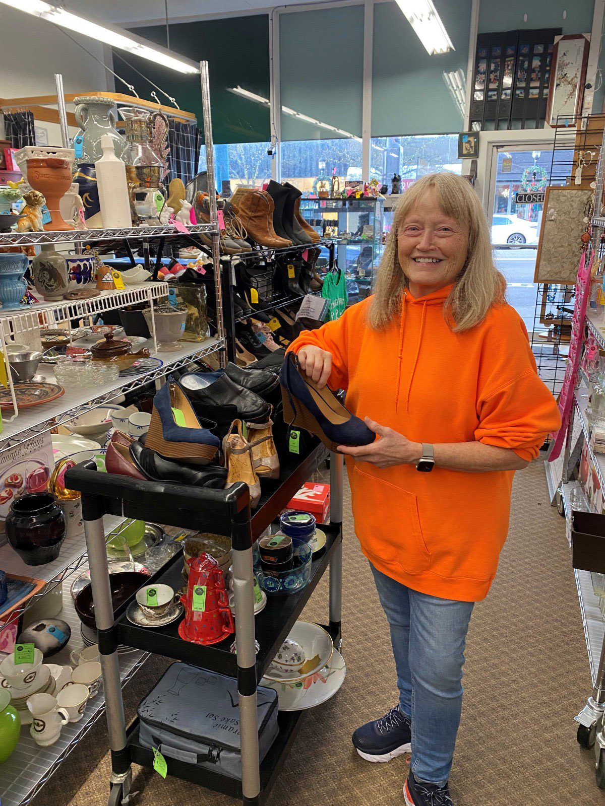 A woman in an orange sweatshirt stands in a secondhand shop, smiling and holding a shoe near shelves filled with various items like dishes, shoes, and decor.