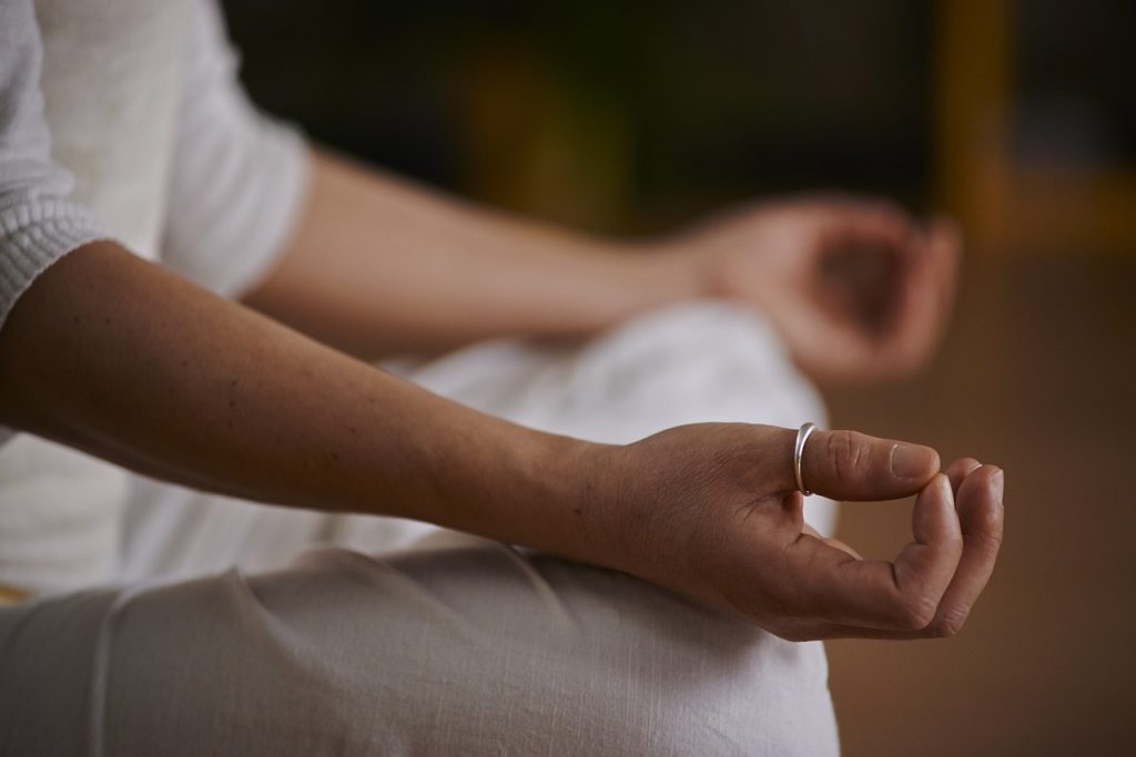 A person wearing white clothes sits cross-legged, with their hand resting on their knee in a meditation pose
