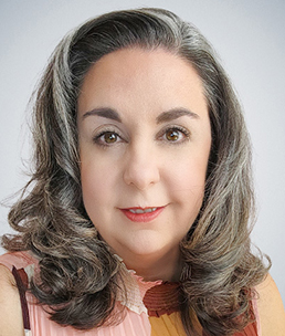 Headshot of Nichole with wavy gray hair and light skin, wearing a multi-colored top, looking directly at the camera against a light background