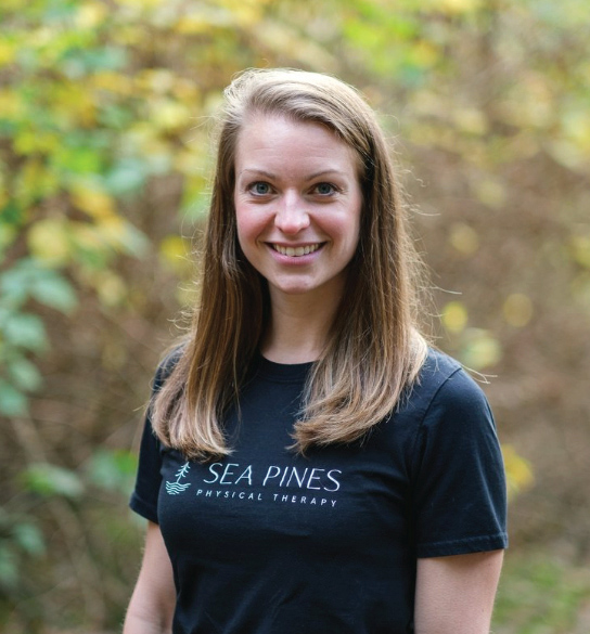 Julia with straight, light brown hair wearing a black “Sea Pines Physical Therapy” t-shirt stands outdoors in front of blurred greenery, smiling at the camera