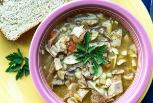 Bowl of soup with noodles, mushrooms, and herbs, garnished with parsley, next to a slice of bread on a yellow plate